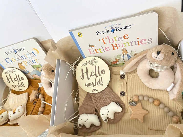 New baby hampers for a baby boy or girl, including a beige romper, plush animal rattle, silicone pacifier clip, hello world plaque and a Beatrix Potter Peter Rabbit storybook.