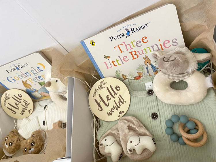 New baby hampers for a baby boy or girl, including a beige romper, plush animal rattle, silicone pacifier clip, hello world plaque and a Beatrix Potter Peter Rabbit storybook.