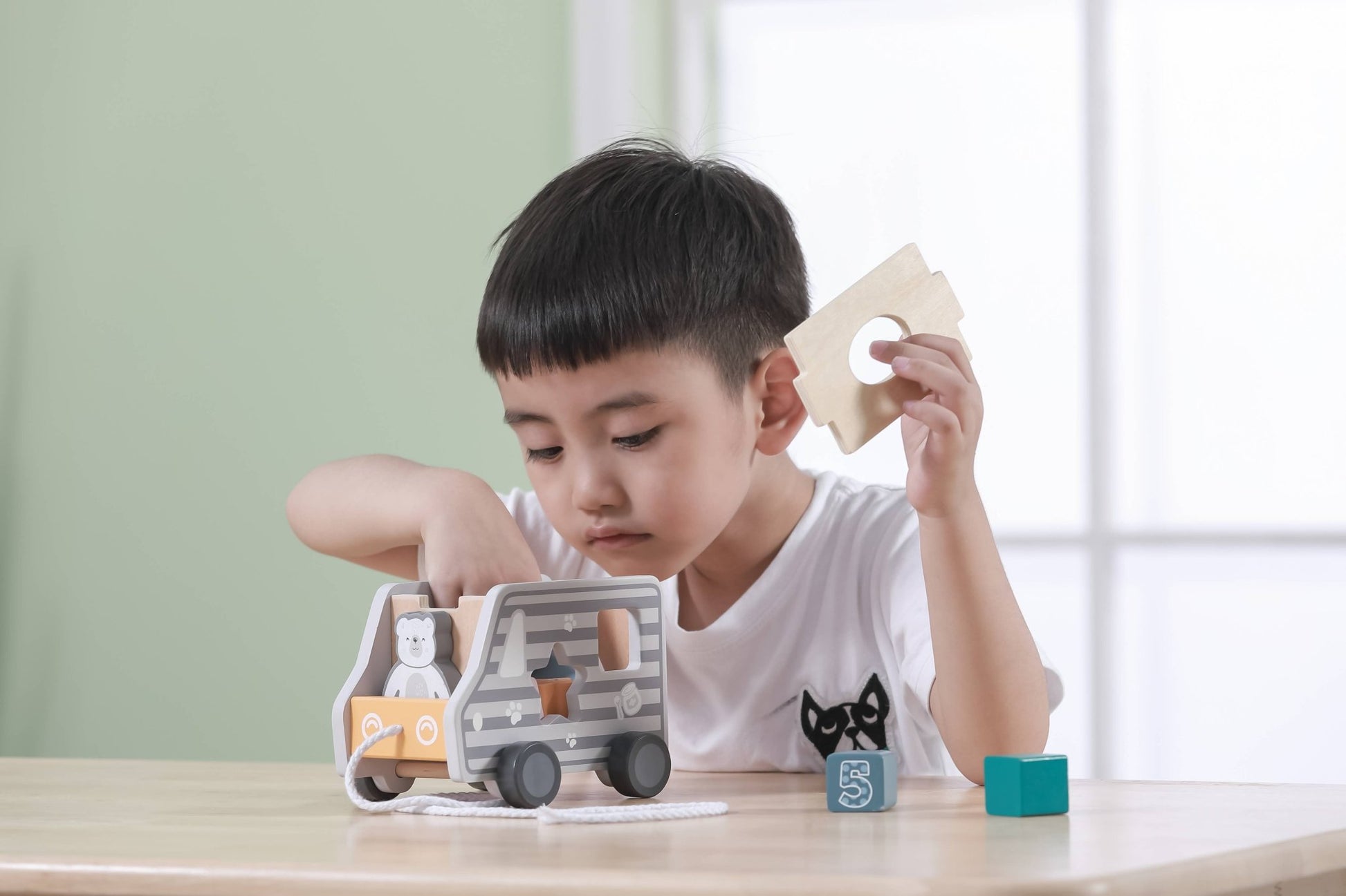 A wooden toy shape sorter made by PolarB designed as a pull-along truck. The truck features a cute bear illustration on the front and gray stripes along the sides. Various numbered blocks, including a star, triangle, and cylinder, are placed around the truck, each with a corresponding shape slot on the side for sorting. A string is attached to the front for pulling the toy.