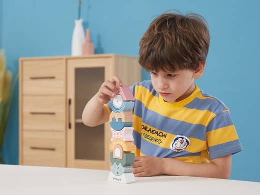 A child playing with a wooden block tower toy puzzle made by PolarB, featuring uniquely shaped and brightly colored pieces stacked vertically. The blocks include various geometric and abstract shapes, such as arches, waves, and a triangle at the top with a smiling bear face. The base of the tower is labeled ‘PolarB.