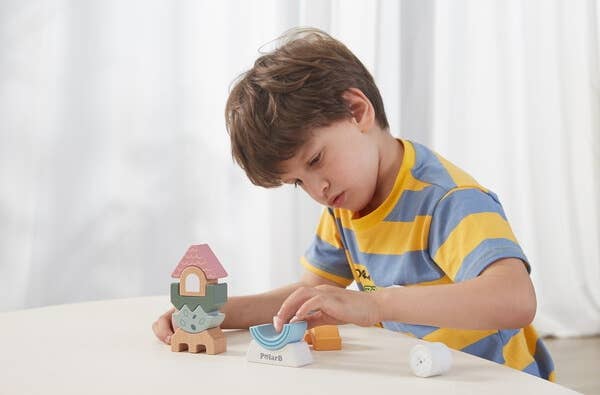 A child playing with a wooden block tower toy puzzle made by PolarB, featuring uniquely shaped and brightly colored pieces stacked vertically. The blocks include various geometric and abstract shapes, such as arches, waves, and a triangle at the top with a smiling bear face. The base of the tower is labeled ‘PolarB.