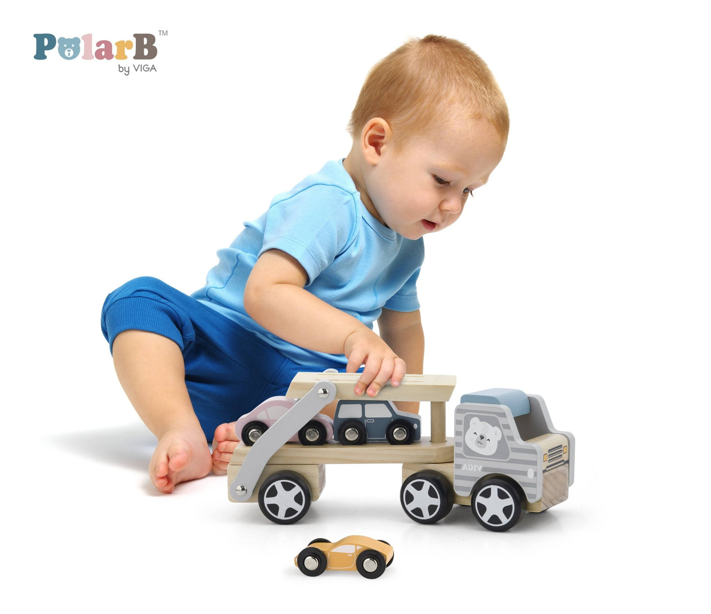 A child playing with a wooden toy car transporter made by PolarB, featuring a light brown carrier truck with a gray-striped cab, an illustrated bear on the side, and a movable two-level ramp. The set includes three small wooden toy cars in pastel yellow, pink, and blue, designed to be loaded onto the carrier.