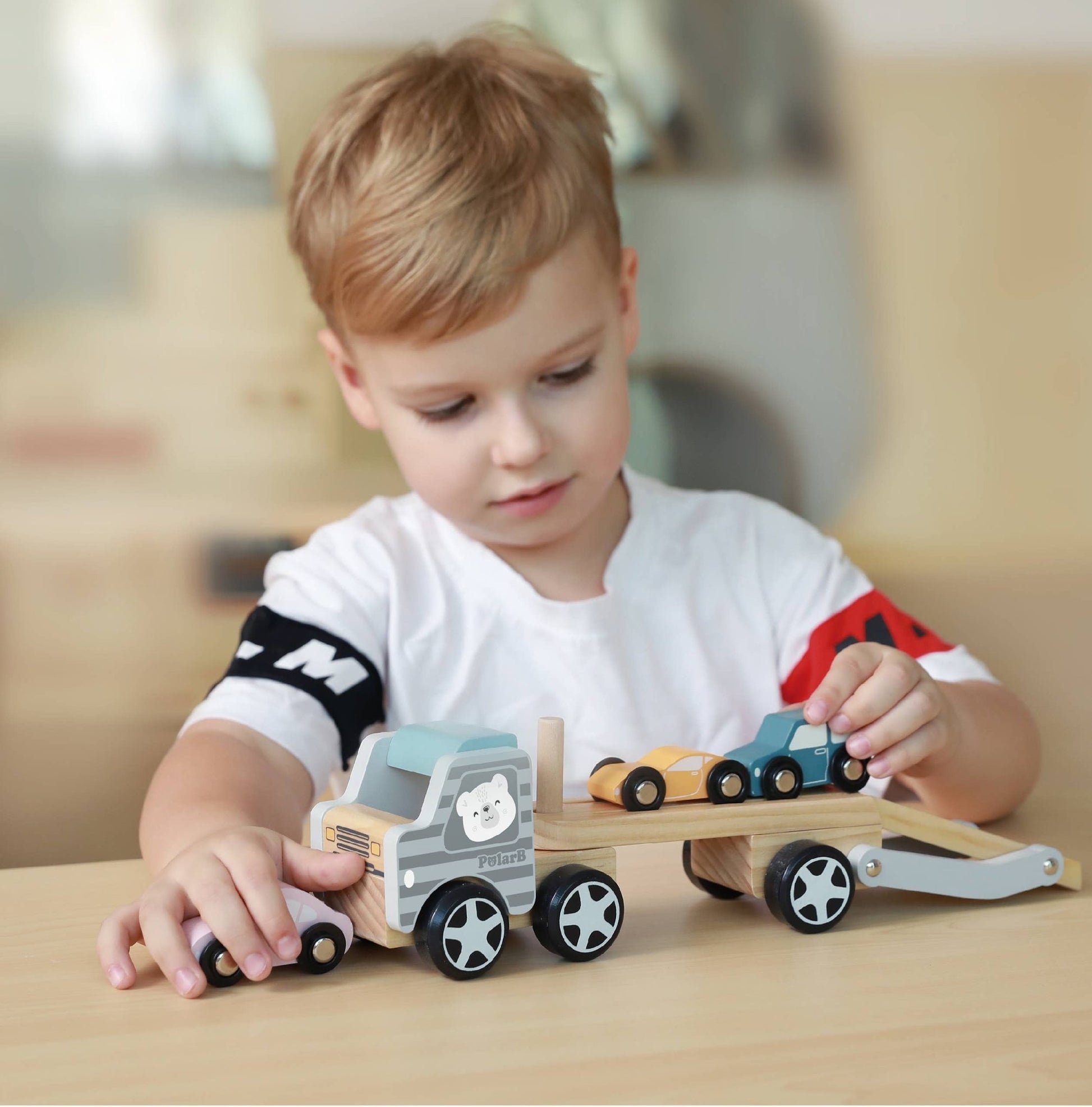 A child playing with a wooden toy car transporter made by PolarB, featuring a light brown carrier truck with a gray-striped cab, an illustrated bear on the side, and a movable two-level ramp. The set includes three small wooden toy cars in pastel yellow, pink, and blue, designed to be loaded onto the carrier.