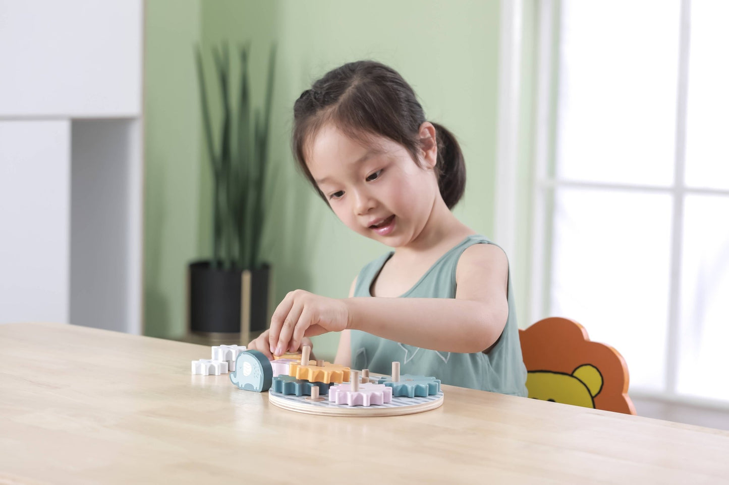 A wooden toy gear puzzle made by PolarB, featuring a circular base with colorful interlocking gears in shades of blue, yellow, pink, and white. The puzzle includes animal figures, such as a blue elephant, pink fox and an orange lion, which can be placed on top of the gears.